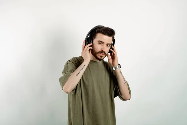 Young man wearing khaki t-shirt posing isolated over white background wireless headphones meditating listening audio guide. Touching to headphone.
