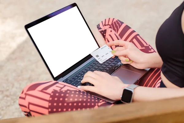 Young caucasian woman wearing sports bra standing on city park, outdoors hands on keyboard and holding credit or debit card. Online shopping, business concept.