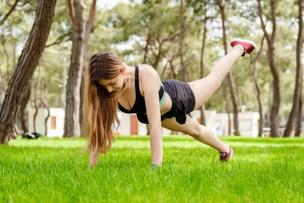 Young beautiful woman wearing sportive clothes on city park, outdoors woman doing push ups with one leg up. Outdoor sports, healthy life concepts.