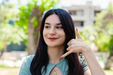 Turkuaz tişört giyen güzel esmer kadın şehir parkında dikiliyor, açık havada gülümsüyor telefonla el ve parmaklarıyla telefonla konuşuyor gibi yapıyor. İletişim kavramları.