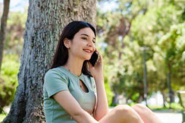 Kentin parkında turkuaz tişört ve turuncu kısa tişört giyen, cep telefonuyla arkadaşlarıyla konuşan ya da gülümseyen erkek arkadaşıyla konuşan genç bir kadın..