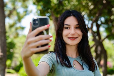 Genç esmer kız turkuaz tişört giyip mutlu bir şekilde gülümsüyor. Şehir parkında dikiliyor, açık havada akıllı telefonuyla selfie çekiyor. Ekrana bakıyor ve selfie çekiyor. Yüzüne odaklan..