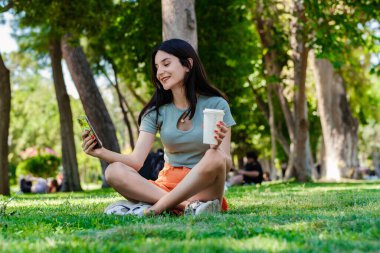 Şehir parkında tişört giyen, elinde kahve fincanı tutan ve cep telefonu kullanan beyaz bir kadın. Açık hava sporları, sağlıklı yaşam kavramları..