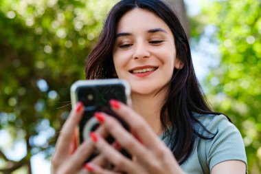 Genç esmer kız turkuaz tişört giyip mutlu bir şekilde gülümsüyor. Şehir parkında duruyor, açık havada telefon ekranına bakıyor ve telefon kullanıyor. Arkadaşlarla mesajlaşma, video izleme veya sosyal medyada kaydırma.