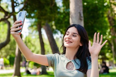 Kentin parkında turkuaz tişört ve turuncu kısa tişört giyerek gülümseyen genç bir kadın, açık havada elinde telefonla ekranda el sallayan el sallama videosu ve mobil uygulama üzerinden uzaktan arama yapan arkadaşına bakıyor..