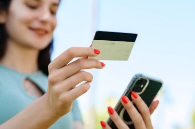 Şehir parkında tişört giyen mutlu bir kadın, elinde cep telefonu kartı olan bir kadın internet üzerinden alışveriş yapıyor ve internet üzerinden ödeme yapıyor. Güvenli mobil bankacılık.