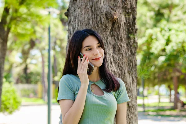 Portrait of brunette woman wearing turquoise tee and orange short on city park, outdoors talking on mobile phone with friends or boyfriend with smiles.