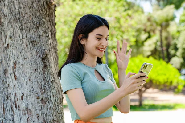 Happy woman wearing tee standing on city park, outdoors holding phone looking at screen waving hand video calling distance friend online in mobile app using smartphone video chat application.