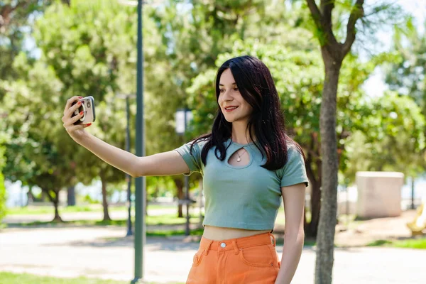 Brunette millennial woman wearing turquoise tee on city park, outdoors taking a self portrait with smart phone. She looks at the screen and taking selfie.