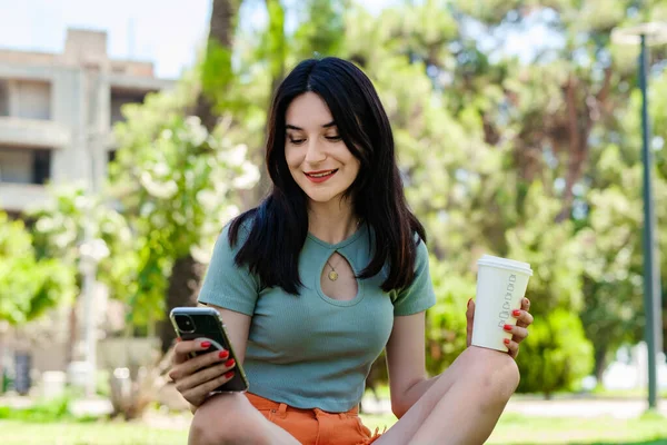 Happy brunette woman wearing turquoise tee on city park, outdoors holding takeaway coffee mug and using mobile smart phone. Scrolling on social media or messaging with friends on cell phone.