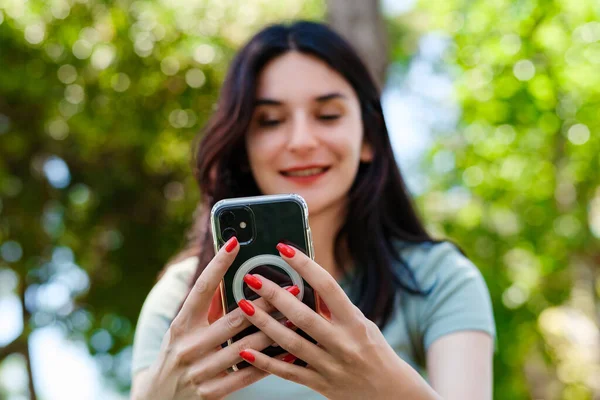 Brunette millennial woman wearing turquoise tee on city park, outdoors looking at the phone screen and using phone. Messaging with friends, watching video or scrolling on social media.