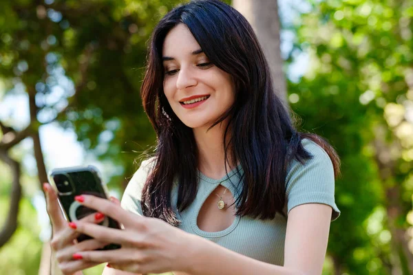 Young brunette woman wearing turquoise tee and orange short on city park, outdoors looking at the phone screen and using phone. Messaging with friends, watching video or scrolling on social media.