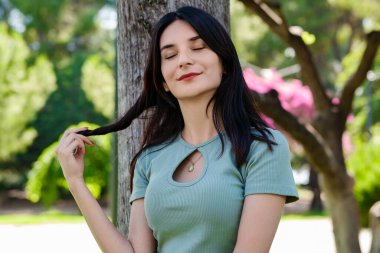 Turkuaz tişört giyen, turuncu kısa tişörtlü, açık havada saçlara dokunan ve kapalı gözlerle düşünen genç esmer kadın. Rüya gibi güzel kadın gülüşü.