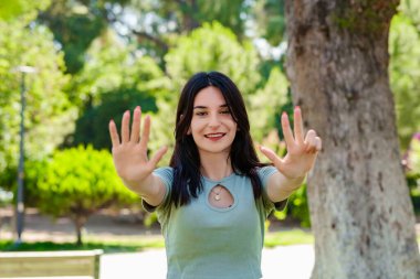 Şehir parkında tişört giyen neşeli esmer kadın sekiz numarayı gösterip gösterirken kendinden emin ve mutlu bir şekilde gülümsüyor. Yüzüne odaklan..