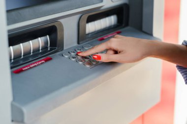 Young brunette woman wearing blue striped shirt on city park, outdoors using ATM machine to withdraw her money.