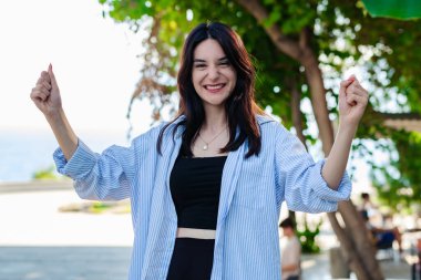 Kentin parkında kendine güvenen, çizgili tişört giyen genç bir kadın mutlu ve heyecanlı bir şekilde kollarını kaldırarak zafer jesti yapıyor ve başarıya gülümsüyor. Zafer hissediyorum..