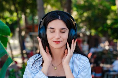 Beautiful brunette woman wearing striped shirt standing on city park, outdoors listening music in headphones and eyes closed. Enjoy good quality sound, stress free concept.