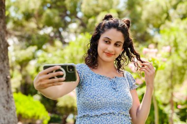 Şehir parkında duran, yazlık elbise giyen mutlu bir kadın akıllı telefonuyla kendi portresini çekiyor. Ekrana bakıyor ve selfie çekiyor..