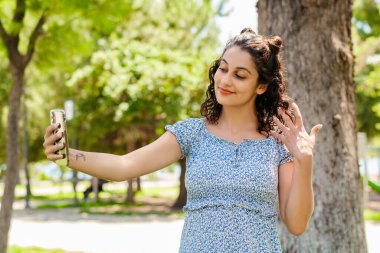 Şehir parkında duran mavi çiçekli bir elbise giyen güzel esmer kadın portresi. Akıllı telefonuyla kendi portresini çekiyor. Ekrana bakıyor ve selfie çekiyor..