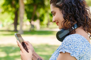 Young woman smiling confident wearing summer dress standing on city park, outdoors looking at the phone screen and using phone. Messaging with friends, watching video or scrolling on social media.