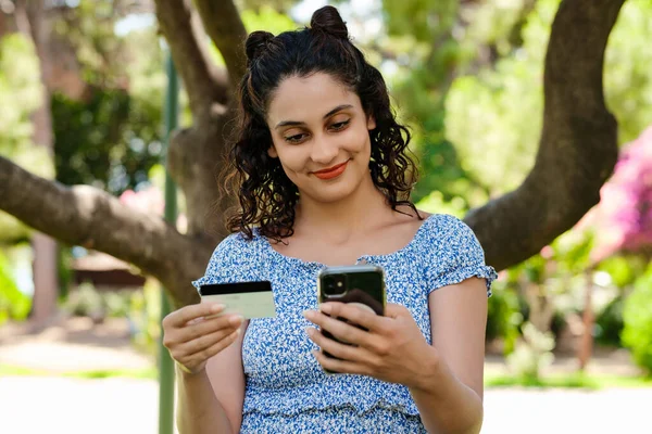 Young brunette girl smiling happy wearing summer dress on city park, outdoors holding mobile phone credit bank card do online shopping provide internet payment. Safe mobile banking.