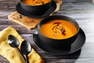 Kabak çekirdeği yağlı turuncu kabak kremalı iki kase, ahşap bir yüzeyde iki siyah kaşık.