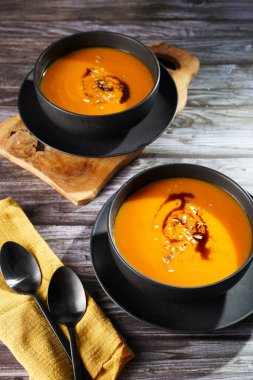 Kabak çekirdeği yağlı turuncu kabak kremalı iki kase, ahşap bir yüzeyde iki siyah kaşık.
