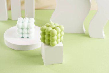 Beyaz küpte yeşil kare bir mum ve arkasında açık mavi bir mum.