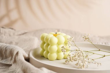 Beton bir tepside, keten bir masa örtüsünün üzerinde, çiçek açan bir dalı olan beyaz kare bir mum.