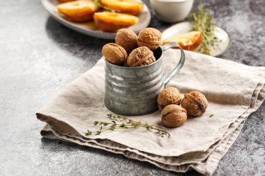 Gri beton bir yüzey üzerinde bej renkli keten kumaş, karamelize armut ve kekik dalları olan kahverengi cevizli metal bir kupa.