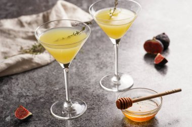 Sarı alkolsüz limonata ve kekik dalıyla iki martini bardağı, bal ve gri mermer yüzeyde incir.
