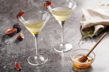 İki martini bardağı, alkollü içki, kekik dalı, bal ve gri mermer yüzey üzerinde incir.