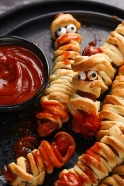 Tatlı Cadılar Bayramı partisi atıştırmalığı: şeker gözlü sosis hamur şeritlerine sarılmış siyah tabakta ketçaplı mumyaları andıran domates sosisi.