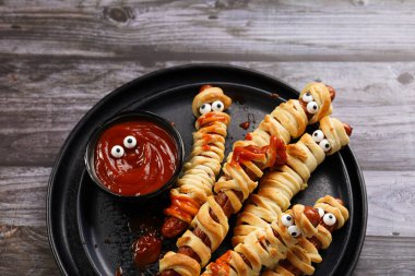 Tatlı Cadılar Bayramı partisi atıştırmalığı: şeker gözlü sosis hamur şeritlerine sarılmış siyah tabakta ketçaplı mumyaları andıran domates sosisi.