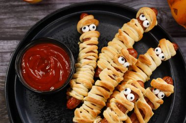 Tatlı Cadılar Bayramı partisi atıştırmalığı: şeker gözlü sosis hamur şeritlerine sarılmış siyah tabakta ketçaplı mumyaları andıran domates sosisi.