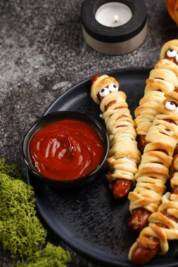 Tatlı Cadılar Bayramı partisi atıştırmalığı: şeker gözlü sosis hamur şeritlerine sarılmış siyah tabakta ketçaplı mumyaları andıran domates sosisi.