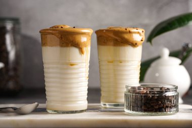 İki bardak, modaya uygun Kore içeceği dalgona kahveli latte. Krem şantili kahve, şeker, sıcak su ve mermer bir tahtada süt katmanları, gri yüzey.