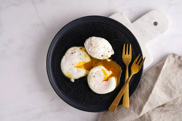 Three poached eggs with egg yolk on a black plate on a marble board and golden forks