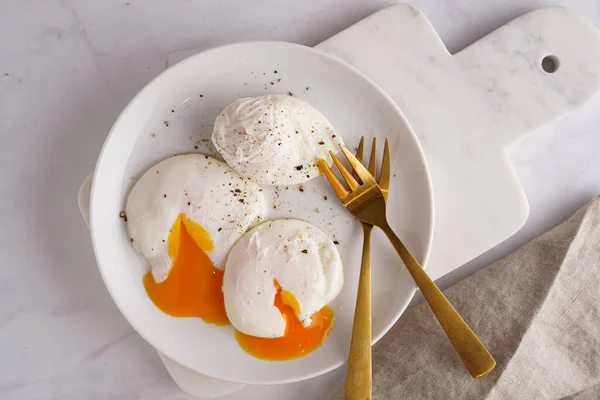 Three poached eggs with egg yolk on a white plate on a marble board and golden forks