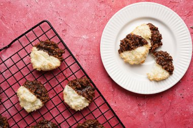 Beyaz bir tabakta çikolataya batırılmış birkaç hindistan cevizi taneli kurabiye ve morumsu yüzeyde fırın rafı.