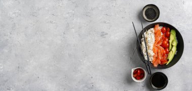 Poke kasesi - beyaz pirinçli siyah kase, tütsülenmiş somon, kiraz domatesleri ve avokado, susam tohumu, soya sosu ve gri beton yüzeyde çubuklar. Üst görünüm, negatif boşluk, pankart boyutu