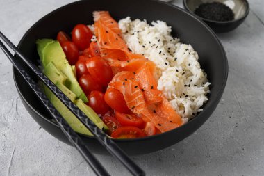 Poke kasesi - beyaz pirinçli siyah kase, tütsülenmiş somon, kiraz domatesleri ve avokado, susam tohumu, soya sosu ve gri beton yüzeyde çubuklar.