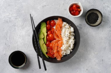Poke kasesi - beyaz pirinçli siyah kase, tütsülenmiş somon, kiraz domatesleri ve avokado, susam tohumları, soya sosu ve çubuklar gri beton yüzeyde, üst manzara