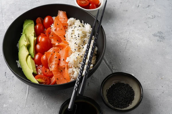 Poke kasesi - beyaz pirinçli siyah kase, tütsülenmiş somon, kiraz domatesleri ve avokado, susam tohumu, soya sosu ve gri beton yüzeyde çubuklar.
