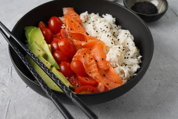 Poke kasesi - beyaz pirinçli siyah kase, tütsülenmiş somon, kiraz domatesleri ve avokado, susam tohumu, soya sosu ve gri beton yüzeyde çubuklar.