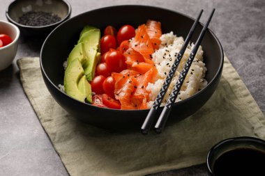 Poke kasesi - beyaz pirinçli siyah kase, tütsülenmiş somon, kiraz domatesleri ve avokado, susam tohumları, soya sosu ve gri beton bir yüzey üzerinde çubuklar, yakın plan.