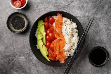 Poke kasesi - beyaz pirinçli siyah kase, tütsülenmiş somon, kiraz domatesleri ve avokado, susam tohumları, soya sosu ve çubuklar gri beton yüzeyde, üst manzara
