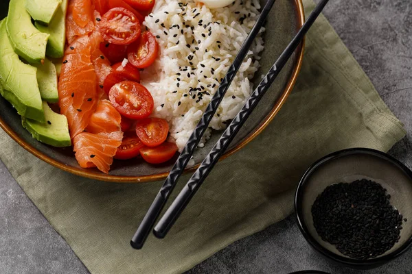 Poke kasesi - beyaz pirinçli yeşil kase, tütsülenmiş somon, kiraz domatesleri ve avokado, susam tohumları, soya sosu ve gri beton bir yüzey üzerinde çubuklar, yakın plan.