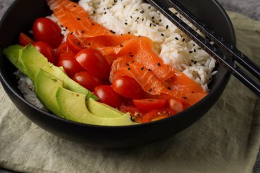 Poke kasesi - beyaz pirinçli siyah kase, tütsülenmiş somon, kiraz domatesleri ve avokado, susam tohumları, soya sosu ve gri beton bir yüzey üzerinde çubuklar, yakın plan.