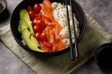 Poke kasesi - beyaz pirinçli siyah kase, tütsülenmiş somon, kiraz domatesleri ve avokado, susam tohumları, soya sosu ve gri beton bir yüzey üzerinde çubuklar, yakın plan.